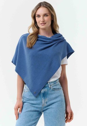 Woman wearing blue triangular shawl over white shirt and light blue jeans, posing against a plain light background.