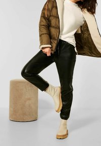 Giacca puffer oliva con fodera crema, maglione bianco a coste, pantaloni in pelle neri e stivaletti beige su uno sgabello cilindrico con texture.