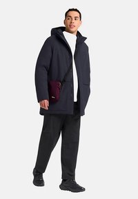 Marineblauwe parka met capuchon, voorzien van ritssluiting en knoopsluiting, voorzakken, gecombineerd met zwarte broek en een kleine bordeauxrode crossbodytas.