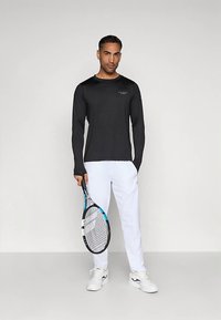 Man står, klädd i en svart långärmad skjorta, vita byxor och vita sneakers, och håller ett svart och blått tennisracket med strängarna vända framåt.