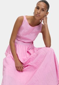 Robe sans manches rose avec un tissu texturé, corsage ajusté et une jupe pleine et fluide. Présente des boutons avec des accents à la taille.