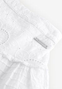 Hvidt bomuldstøj med broderede blomsterdekorationer, blonder og flæset struktur, med et sølvfarvet mærke fra mærket fastgjort.