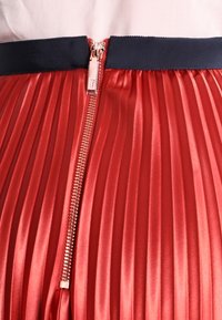 Falda plisada roja con una textura suave y brillante, que presenta una cinturilla de color marino y un cierre de cremallera en la parte posterior con herrajes plateados.