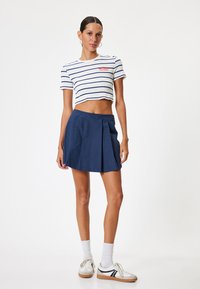 Kurzer blauer Faltenrock, kombiniert mit einem weißen, gestreiften Cropped-Top mit rotem Text. Modell trägt weiße Socken und Sneakers.