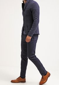 Námořnický blazer z vlněné směsi se dvěma předními kapsami, nošený přes pruhovanou košili, v kombinaci s tmavými kalhotami a hnědými brogue botami.