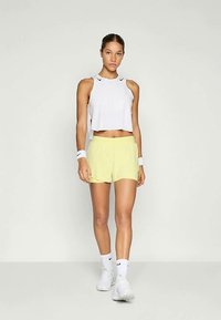 Žena u bijelom bezrukavnom crop topu, žutim šortsevima, bijelim ručnim zavezima, čarapama i tenisicama, stoji protiv jednostavne svijetle pozadine.