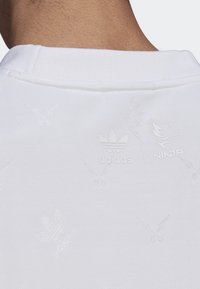 Bílá mikina se subtilním embossovaným vzorem a decentním brandingem s logy "adidas" a "Ninja" poblíž límce.