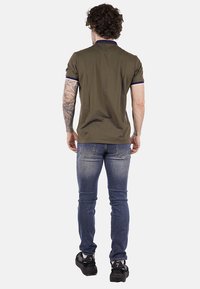 Polo verde oliva con maniche corte, colletto navy a contrasto e jeans slim-fit blu scuro. Il modello indossa sneakers nere.