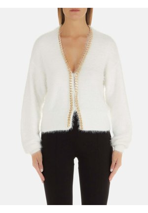 Woman wearing a white fuzzy cardigan with gold chain trim along the deep V-neck and black pants, standing against a white background.