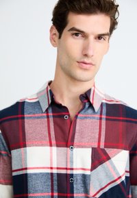 Joven con cabello corto castaño que lleva una camisa de cuadros roja, blanca y azul marino, mirando ligeramente de lado sobre un fondo blanco.