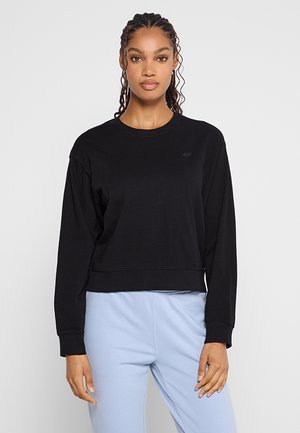 Czarna, krótka bluza z długim rękawem, gładka w dotyku, z okrągłym dekoltem i subtelnym logo na piersi. Zestawiona z jasnoniebieskimi spodniami.