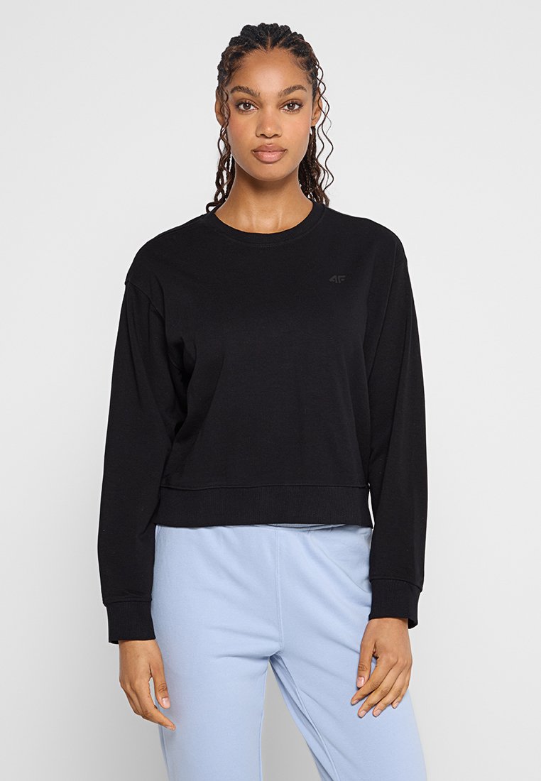 Czarna, krótka bluza z długim rękawem, gładka w dotyku, z okrągłym dekoltem i subtelnym logo na piersi. Zestawiona z jasnoniebieskimi spodniami.