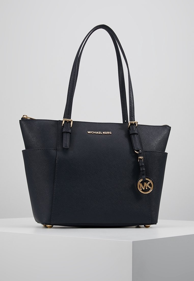 Sac cabas noir Michael Kors avec double anses, plaque logo dorée et breloque ronde MK, posé sur une surface blanche sur fond neutre.