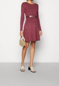 Robe bordeaux à manches longues et côtelée, avec un corsage ajusté et une jupe évasée. Accompagnée d'un sac à main beige et de mules à talons épais de couleur claire.