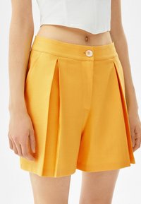 Bermudas amarillas con plisado y una textura suave, que cuentan con un cierre de botón y un diseño de cintura alta. Apta para usar en clima cálido.