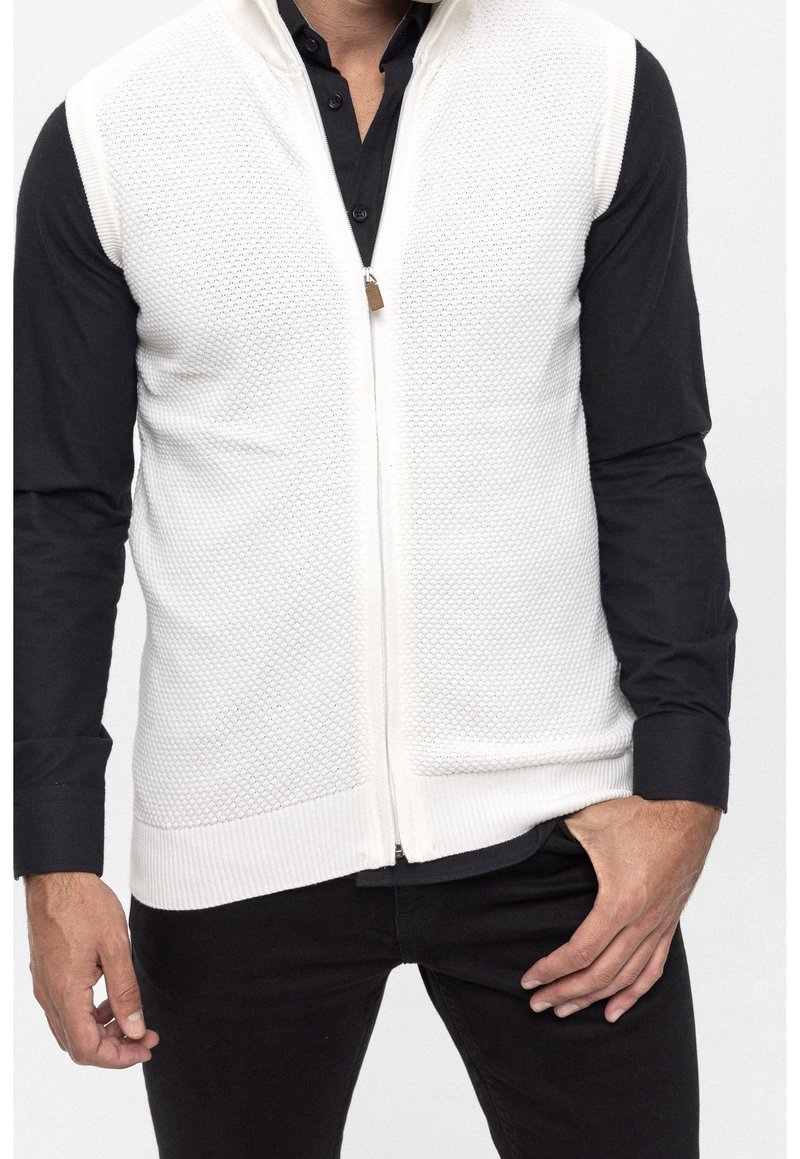 Gilet bianco lavorato a maglia con motivo strutturato, zip frontale e orlo a coste, indossato sopra una maglietta nera a maniche lunghe. Tasche non visibili.