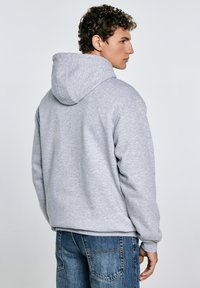 Ein junger Mann mit lockigem Haar trägt einen grauen Kapuzenpullover, von hinten gewandt, kombiniert mit blauen Jeans. Der Hintergrund ist schlicht und hell.