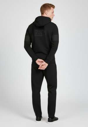 Schwarzes Hoodie- und Hosen-Set mit mattem Logo auf dem Rücken, gerippten Bündchen und klassischer Passform. Der Stoff wirkt glatt und weich.