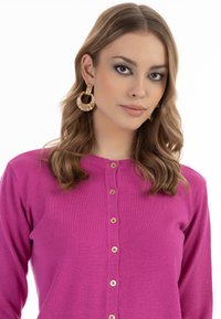Cardigan fucsia con bottoni in legno e tessuto testurizzato, dotato di scollo rotondo. Il modello ha capelli lunghi e mossi e indossa orecchini a cerchio dorati.