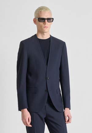 Hombre con cabello rubio corto que lleva gafas de sol negras, chaqueta de traje azul marino oscuro y pantalones a juego, sobre un fondo liso.