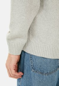 Lichtgrijze gebreide trui met geribbelde manchetten en zoom, gecombineerd met blauwe denim jeans met een achterzak en zichtbare stikseldetails.