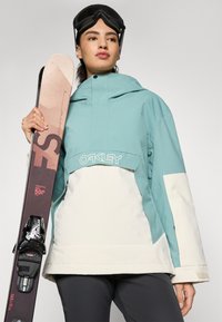 Jasnoniebieska i kremowa kurtka narciarska z wysokim kołnierzem, z logo "Oakley". Model trzyma brązową i czarną nartę z wiązaniami.