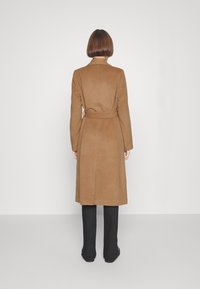 Brauner Trenchcoat mit einem taillierten Gürtel, langen Ärmeln und gerader Silhouette, aus weichem Material. Trägt man über schwarzen Hosen.