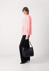 Rosa Langarmshirt mit lockerer Passform, kombiniert mit schwarzen weit geschnittenen Hosen mit subtilen Verzierungen. Das Model hält eine schwarze Lederhandtasche.