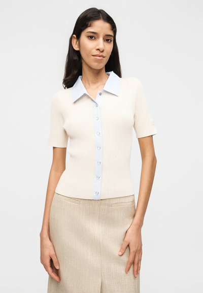 Γυναίκα με μπεζ ριμπ πλεκτό τοπ με κοντά μανίκια και κουμπιά, με ανοιχτό μπλε ριγέ γιακά και μπεζ φούστα, μπροστά σε απλό λευκό φόντο.