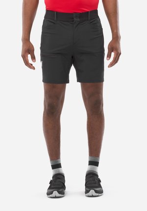 Homme portant un short de sport noir, des chaussettes grises et noires, et des baskets noires, debout devant un fond blanc.