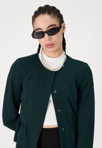 Dark green peplum jacket with a round neckline, black buttons, paired with a white ribbed top and a layered pearl necklace.
