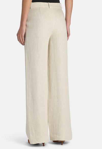Femme portant un pantalon en lin beige taille haute à jambes larges et des chaussures marron à talons, vue de dos sur un fond uni.