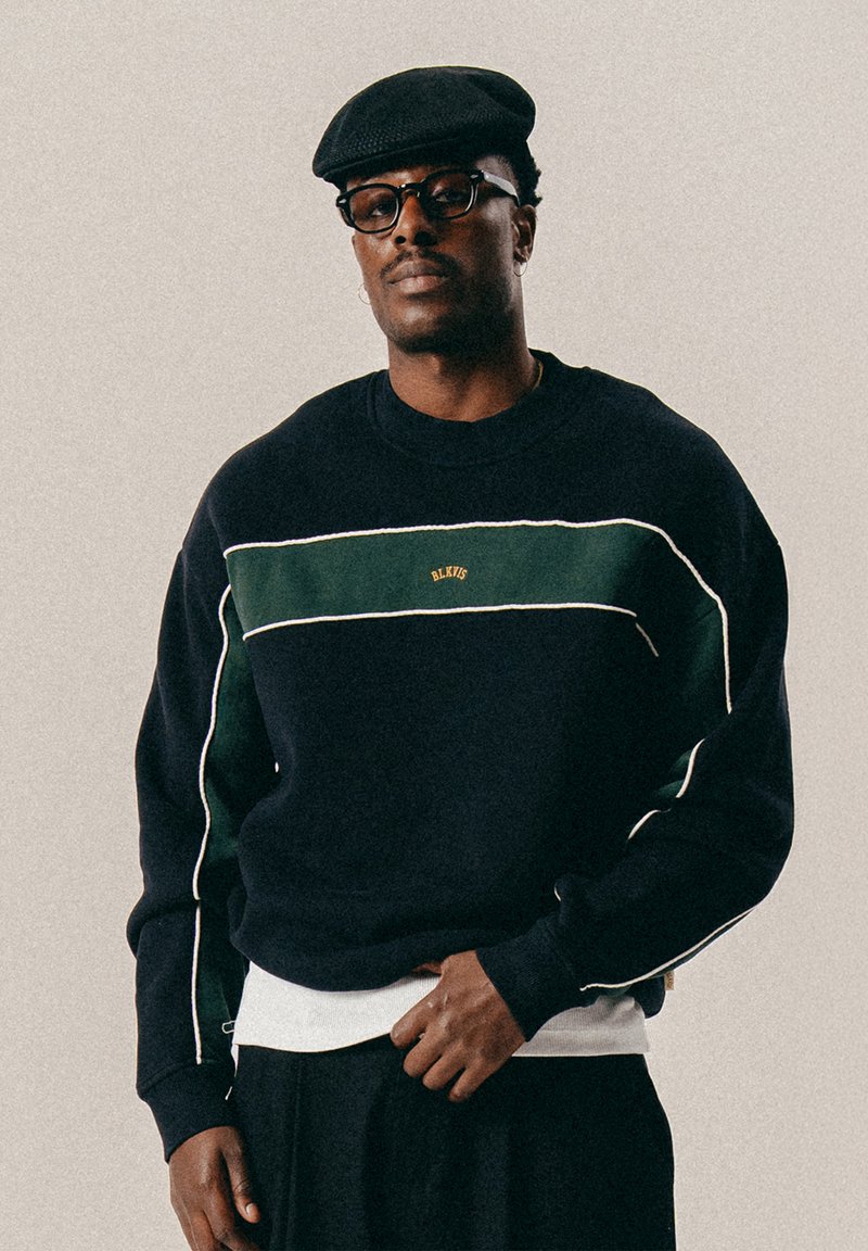 Tumesinine sviiter roheliste triipude, valge äärestuse ja kuldse logoga. Koonilised mansetid ja serv. Kantud valge alussärgi ja musta mütsiga.