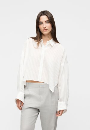 Vrouw met lang bruin haar draagt een oversized cropped blouse met witte krijtstrepen en lichtgrijze, hoogtallige broek, staand tegen een effen achtergrond.