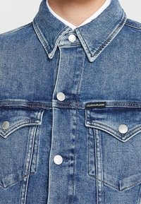 Jeansová bunda v světle modré barvě, s špičatým límečkem, zapínáním na knoflíky vpředu, dvěma kapsami na hrudi s klopami a s označením značky vpředu.