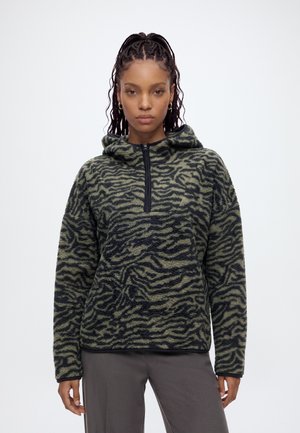 Femme portant un polaire à capuche motif zèbre vert et noir avec une fermeture éclair à l'avant et un pantalon gris, debout devant un fond uni.