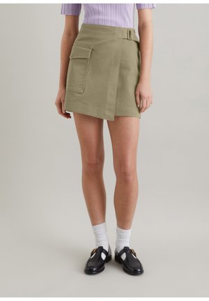 Jupe vert olive avec un design croisé, poche latérale et détail de boucle. À assortir avec des chaussures noires et des chaussettes blanches à la cheville. Tissu texturé.