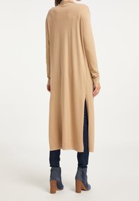 Cardigan long beige à coupe décontractée, avec fentes latérales et manches longues, associé à un jean foncé et des bottines bleues. Texture lisse.