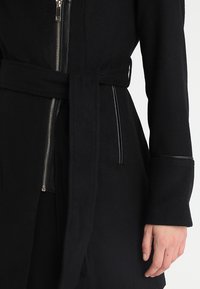 Cappotto di lana nera con un design strutturato. Presenta una zip frontale, vita con cintura e dettagli in pelle sulle maniche e tasche.