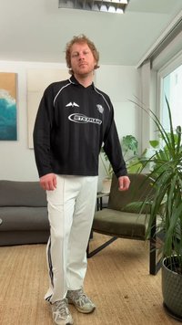 Jersey preto de manga comprida com riscas brancas e logótipo, combinado com calças brancas com riscas laterais escuras, completo com sapatilhas desportivas cinzentas.