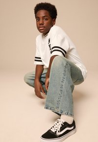Weißes kurzärmliges Hemd mit schwarzen Streifen und "NY"-Druck, hellblaue lässige Jeans und schwarze Sneakers mit weißen Akzenten. Lässige Pose.