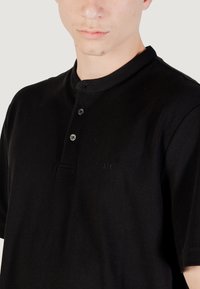 Camisa polo negra de manga corta con una tela texturizada, que cuenta con un cuello redondo, una tapeta con tres botones y un sutil logo en el pecho.