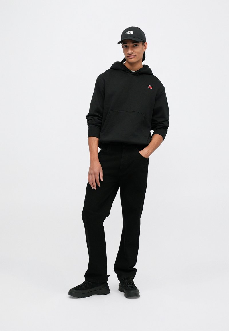 Jeune personne portant un sweat à capuche noir avec un patch rouge, un pantalon noir, des baskets noires et une casquette noire, posant avec une main dans sa poche sur un fond blanc.