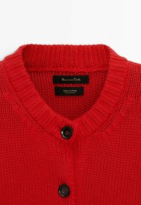 Röd bomullsstickad cardigan med ribbad krage, två svarta knappar vid halsringningen och en mjuk textur med ett öppet vävmönster.