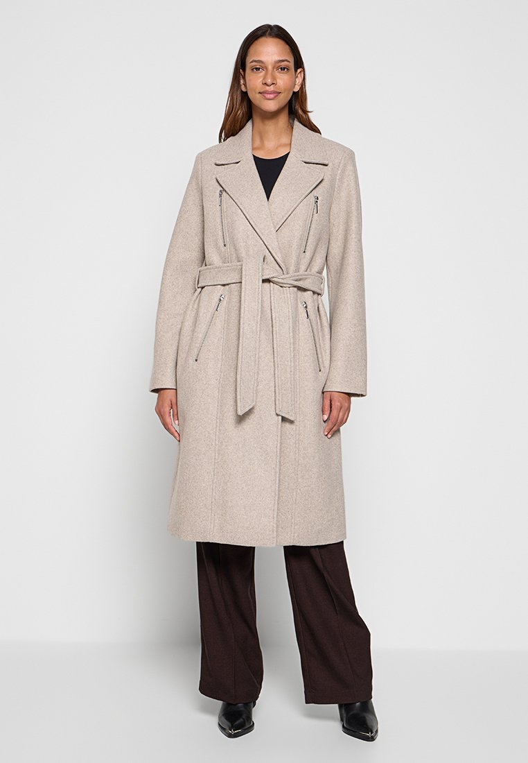 Frau in beigem, tailliertem Wollmantel mit Reißverschluss-Taschen, schwarzem Top, dunkelbraunen weiten Hosen und schwarzen spitzen Schuhen, die vor einer weißen Wand steht.