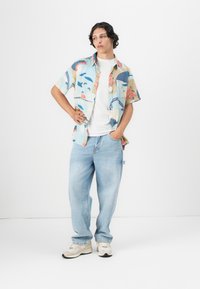 Cămașă ușoară, cu mâneci scurte, cu modele abstracte multicolore; purtată peste un tricou alb. Jeans albastri deschis, cu croială lejeră și adidași.