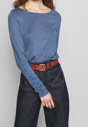 Vrouw die een blauwe lange mouw blouse draagt, ingestopt in een donkere spijkerbroek met witte stiksels en een bruine riem met een gouden gesp.