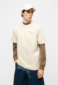 Mladi moški, oblečen v bež Puma majico preko rjave dolge majice, modre kavbojke in ujemajočo se bež kapo, gleda na stran.