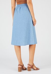 Jupe en denim bleu clair avec un design évasé, une taille élastique et une coupe à mi-genoux. Associée à des sandales à talons bruns. Texture lisse.