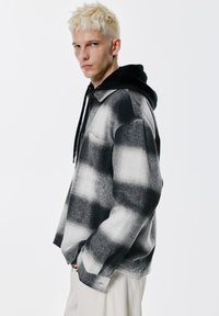 Velika košulja crno-bijele karirane tkanine s ovratnikom, uparena s crnom duksericom s kapuljačom. Mekani materijal, ležerni kroj.